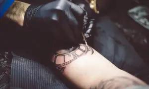 Artysta w czarnej rękawiczce tworzy tatuaż zegara z rzymskimi cyframi, stosując technikę whip shading dla uzyskania głębi.