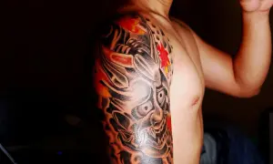 Męskie ramię zdobi tatuaż z demoniczną maską oni i czerwonymi liśćmi klonu.