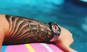 Ramię z realistycznym tatuażem w stylu tribal, ozdobione zegarkiem, spoczywa na dmuchanym materacu przy basenie.