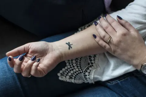 Delikatne, czarne tatuaze male na nadgarstku, w tym ważka. Dłoń ozdobiona ciemnym lakierem i pierścionkiem.