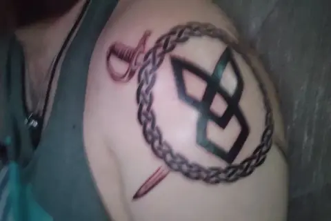 Tatuaż na ramieniu: symbol odrodzenia, spleciony łańcuch, szabla i sztylet.
