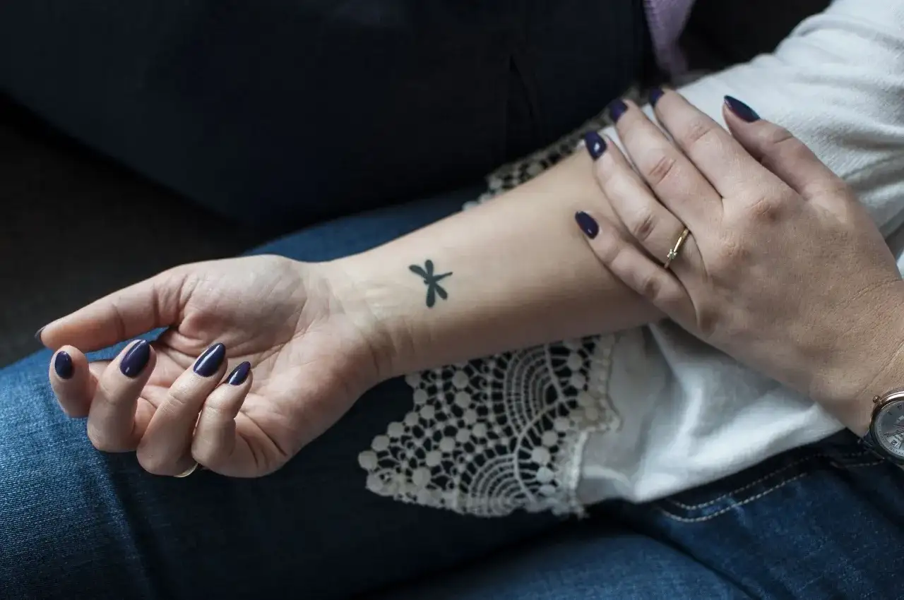 Delikatne, czarne tatuaze male na nadgarstku, w tym ważka. Dłoń ozdobiona ciemnym lakierem i pierścionkiem.